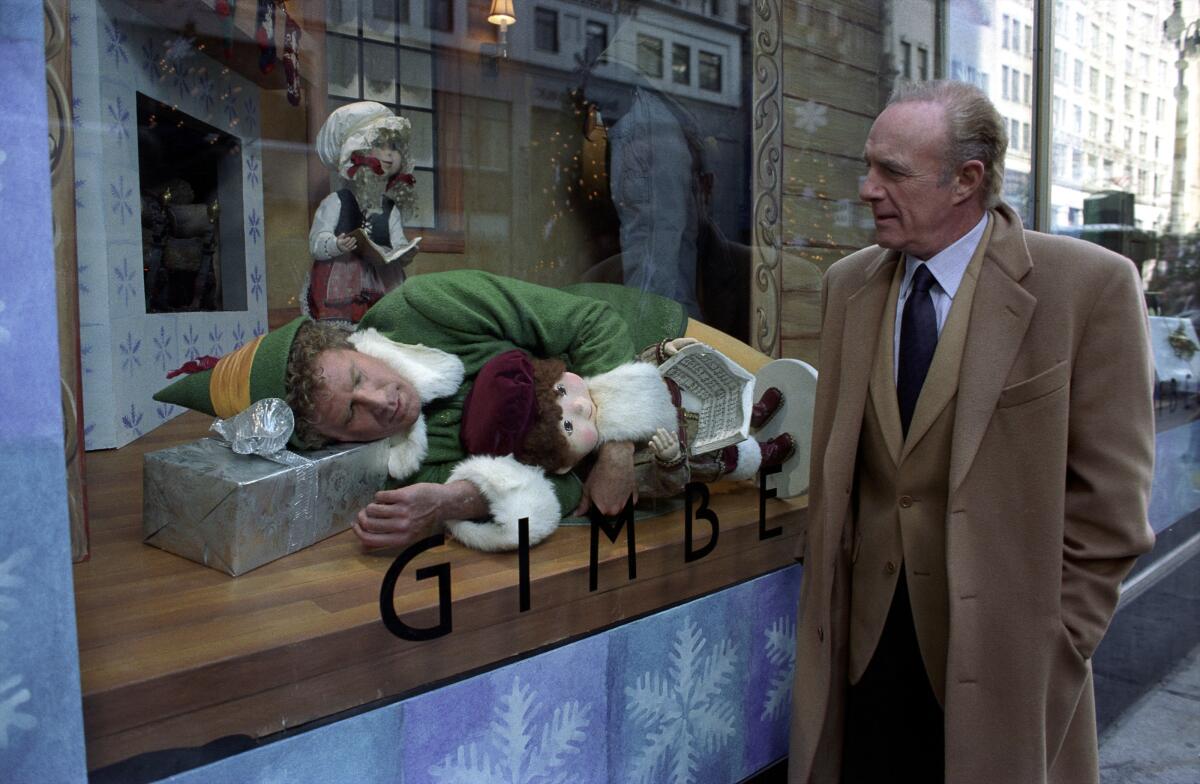 A man dressed as a Christmas elf lies down in a store window as a man in a coat walks by and stares.