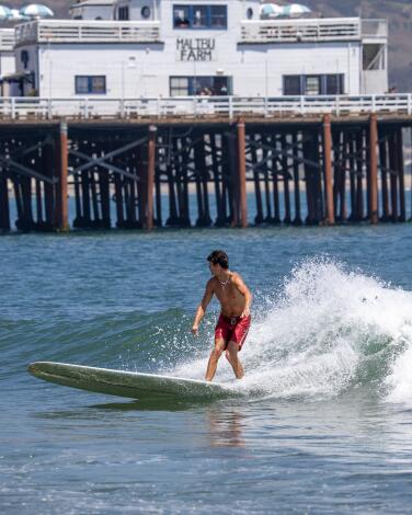 A surfer in red trunks surfs in front of the Malibu pier. 