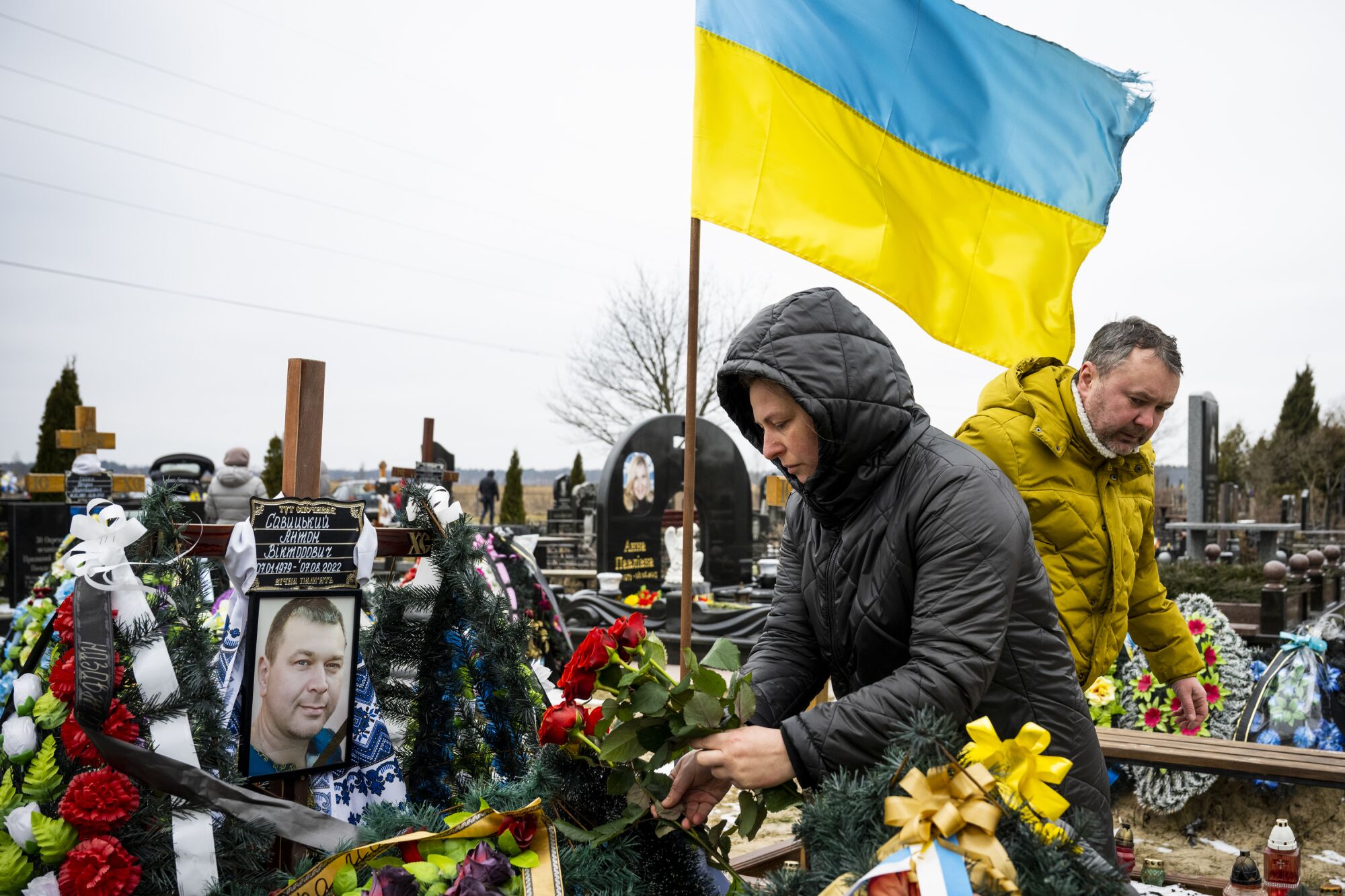 Mourners in raincoats visit a grave adorned with flowers and the Ukrainian flag
