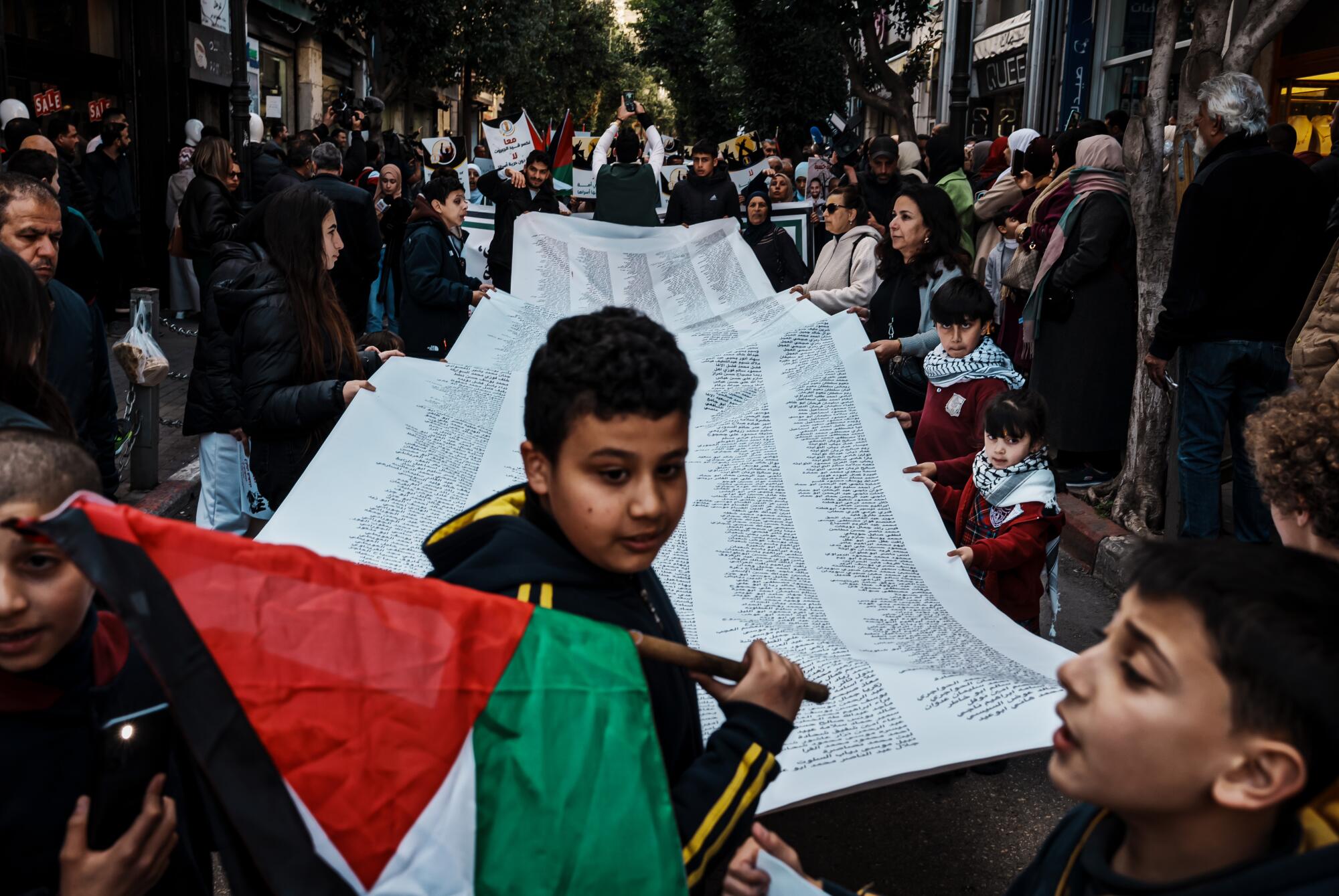 People march and hold a long banner with names of people killed in the war. A child holds a Palestinian flag. 