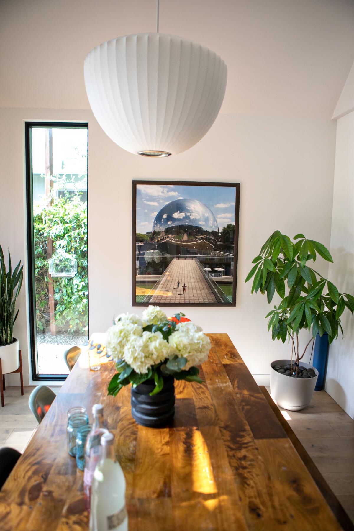 A wooden dining table with flowers on it, a bubble lamp above and a photo on the wall behind