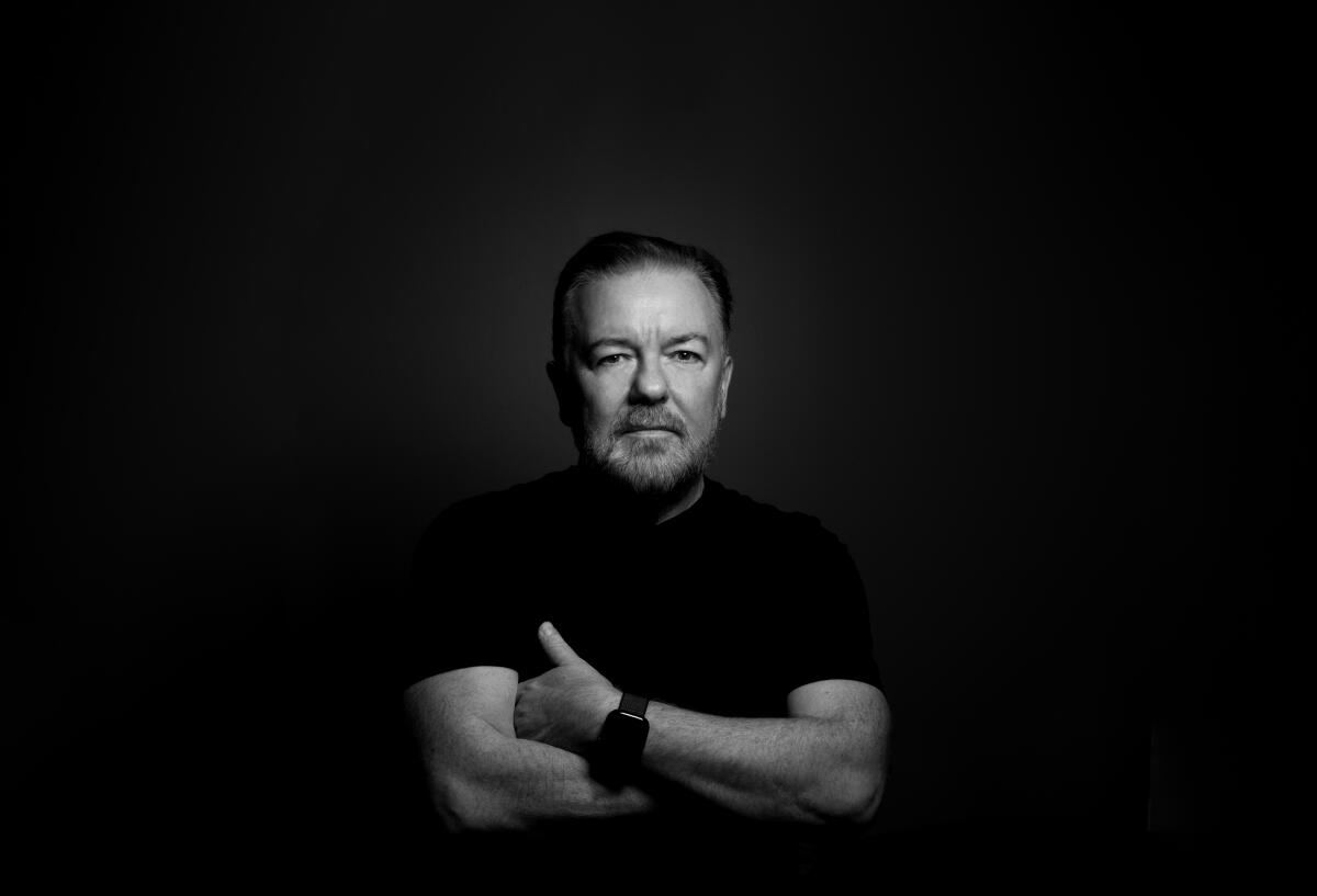 Ricky Gervais in a black shirt stares straight ahead with arms crossed