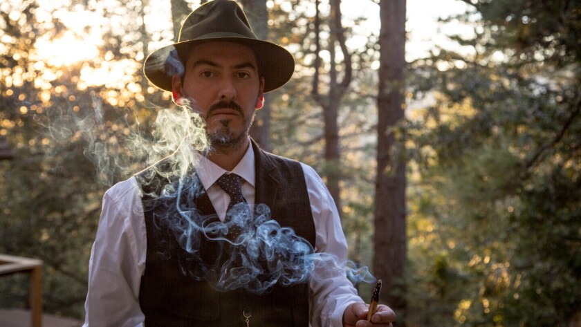 Dhani Harrison wears a hat, vest and tie as a plume of smoke wafts in front of him.