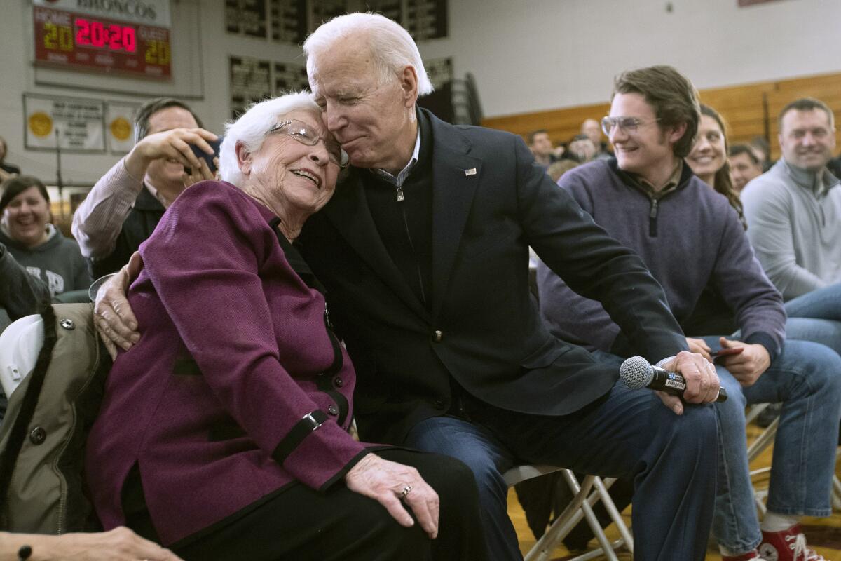 A woman sitting next to Joe Biden in a crowd leans her head on his shoulder as he puts an arm around her
