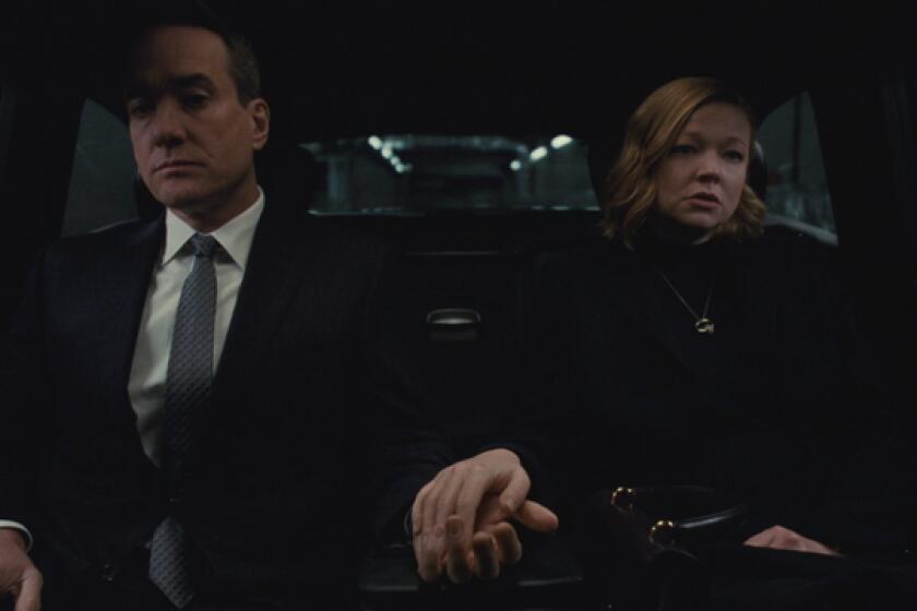 A husband and wife, dressed all in black, hold hands in the back of a car.