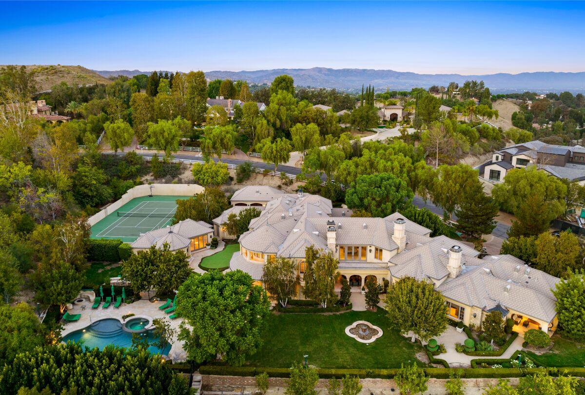 Aerial view of an estate with a large mansion, tennis court and pool surrounded by trees.