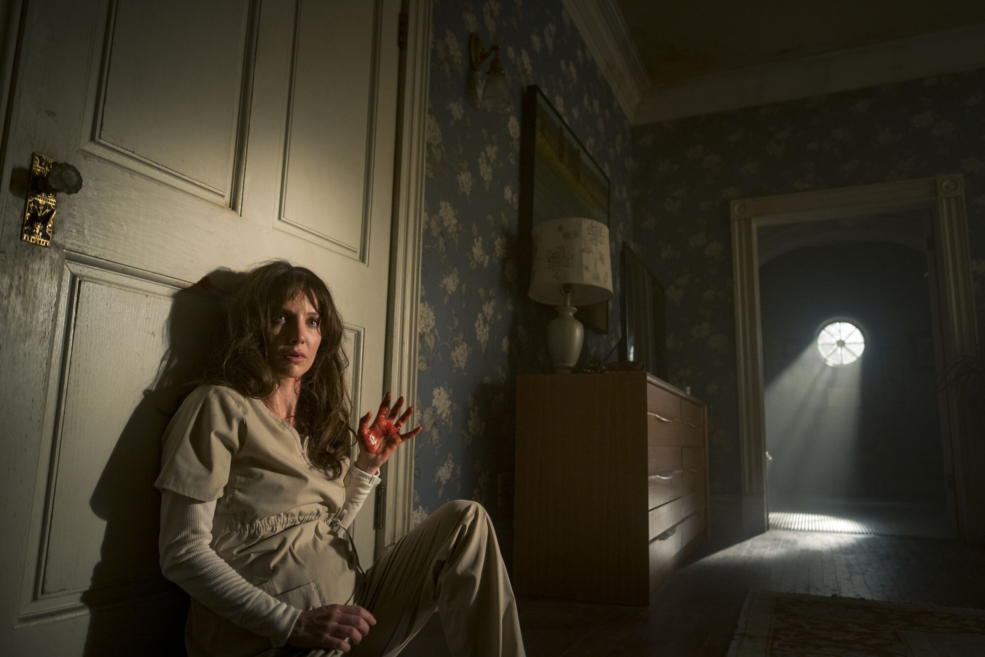 A woman sits on the floor against a closed door in spooky lighting