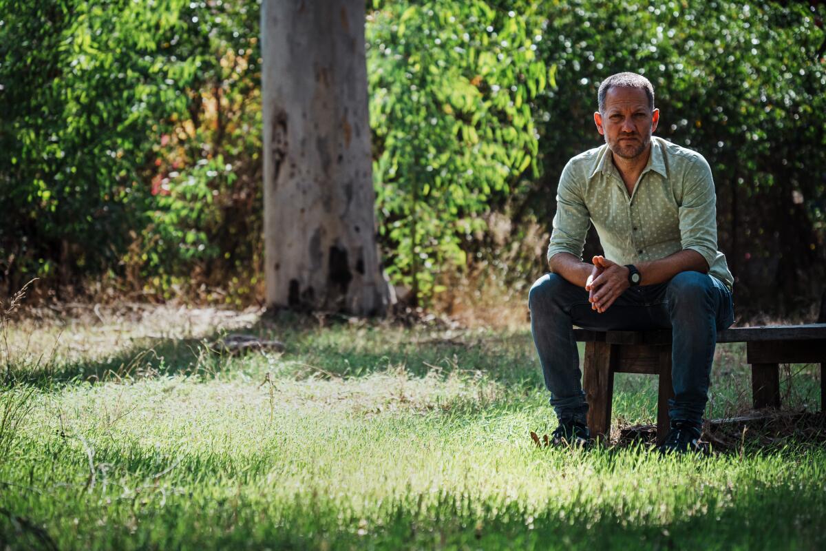 A man sitting, hands together, on a bench in a grassy area shaded by trees