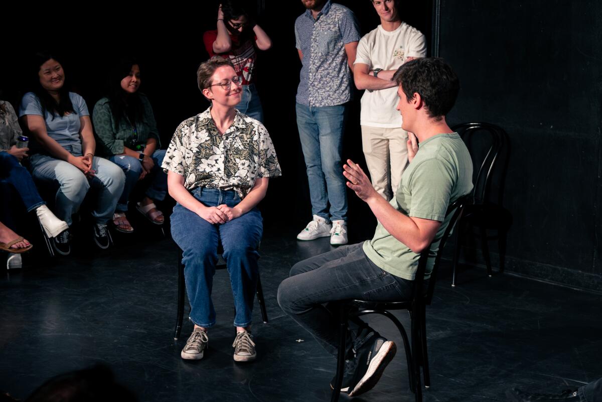 Two people seated onstage as other standing and seated people watch them