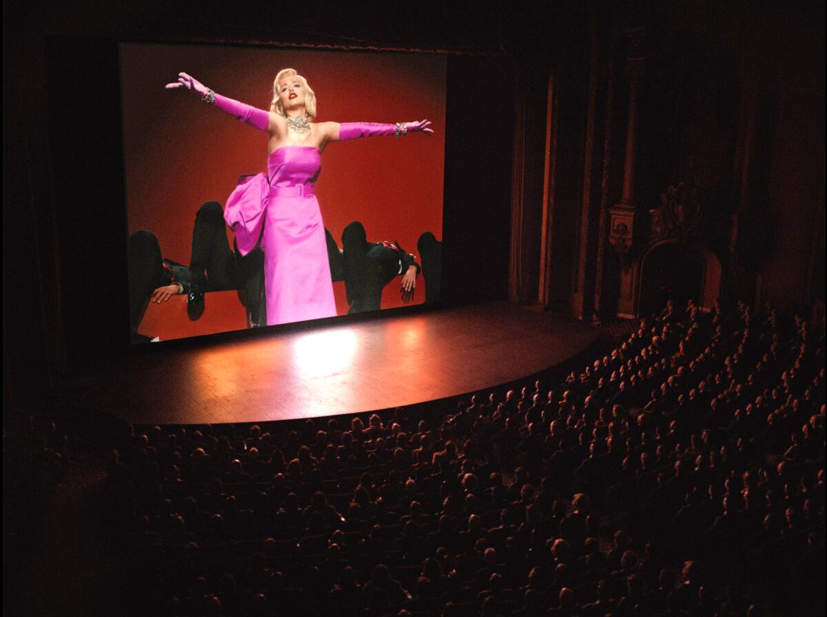 A blond woman in a pink formal dress and jewels is seen on a movie screen.