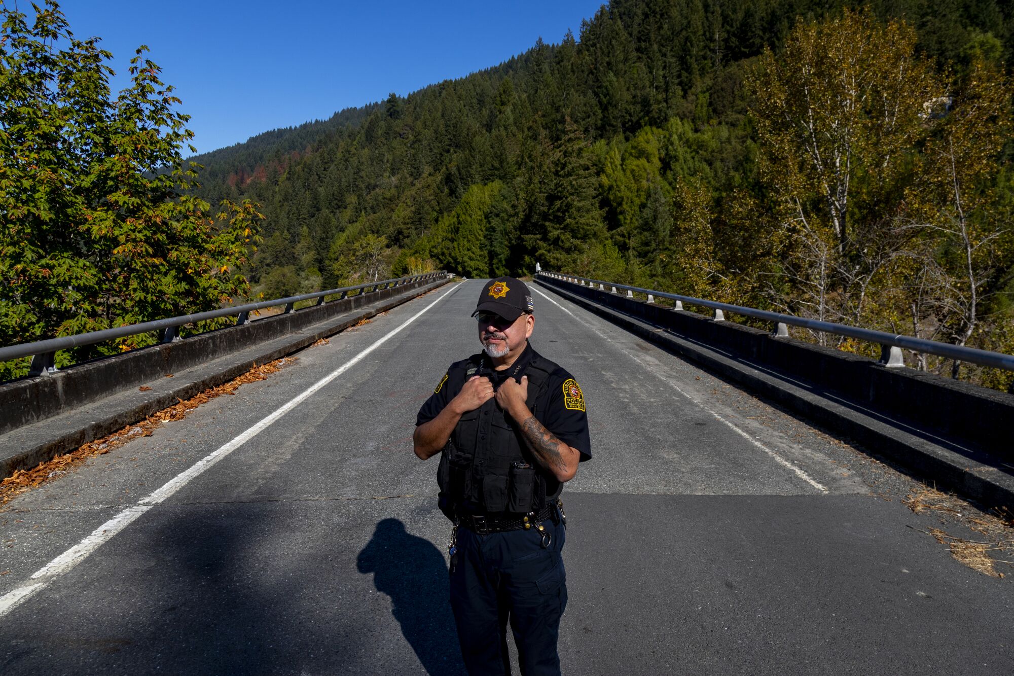 Yurok Tribal Police Chief Greg O'Rourke는 주로 소나무 숲으로 둘러싸인 도로 다리에 서 있습니다.