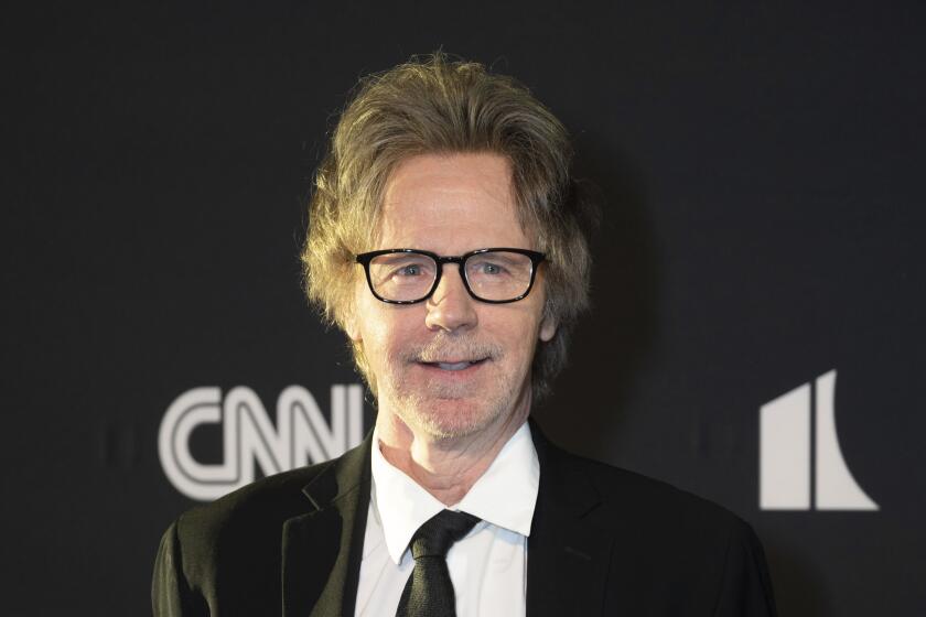 Dana Carvey with thin black glasses and a black suit and tie smiling and posing against a black background