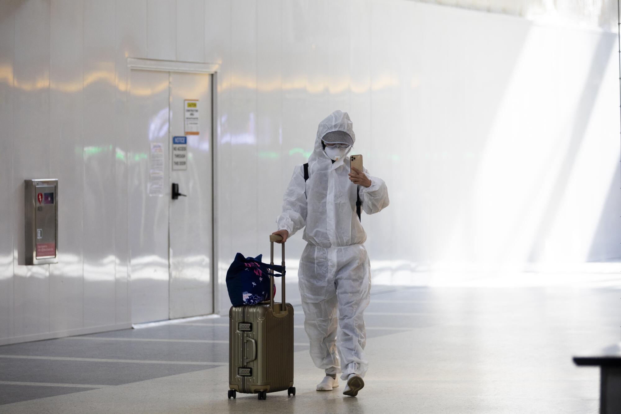 A person wears a full-body protective suit while pushing their luggage through an airport hallway