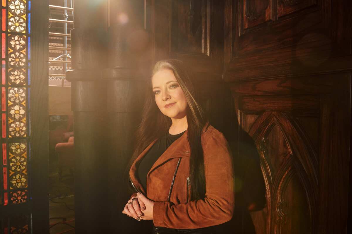 A female country singer in a brown suede jacket stands against a carved wood wall.