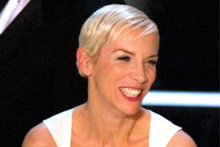 Singer Annie Lennox is wearing a white dress and smiling as she holds her Oscar