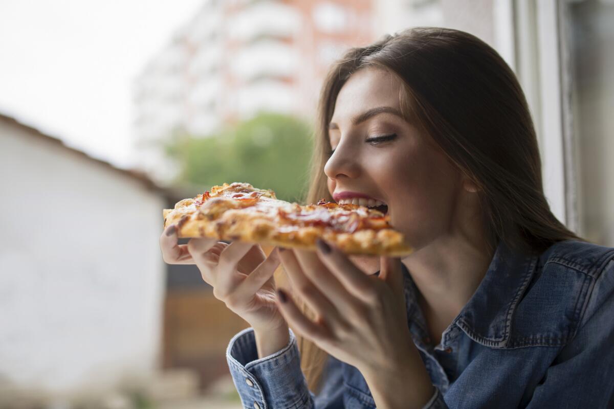 Beautiful, young woman leaning at the window, eating pizza and enjoying
