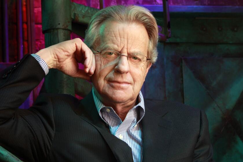 Jerry Springer looks at the camera while leaning on a railing, resting the side of his head on his hand. 