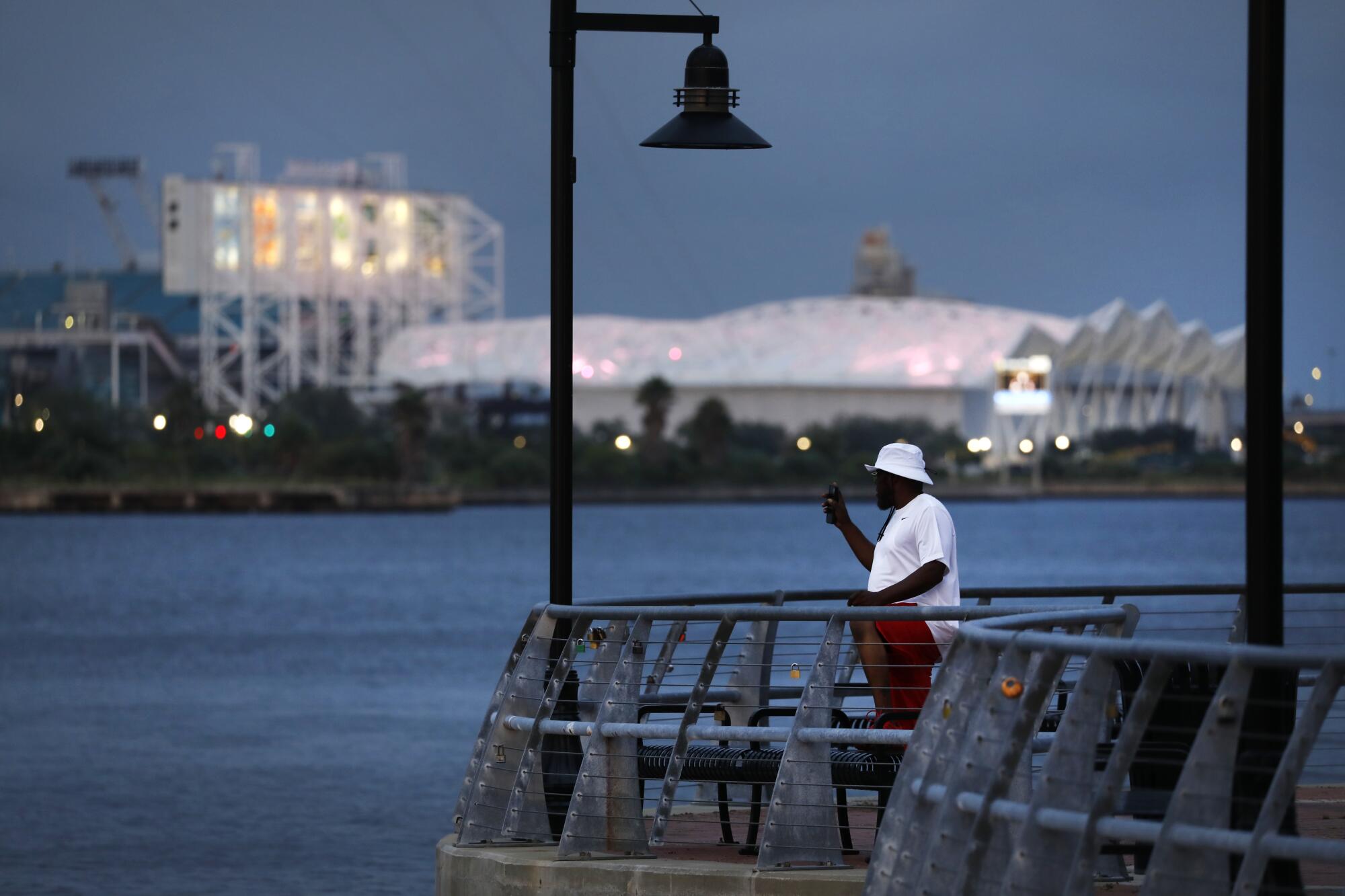 A man in a white shirt and hat and red pants takes pictures along a river, across from a white stadium that is lighted up