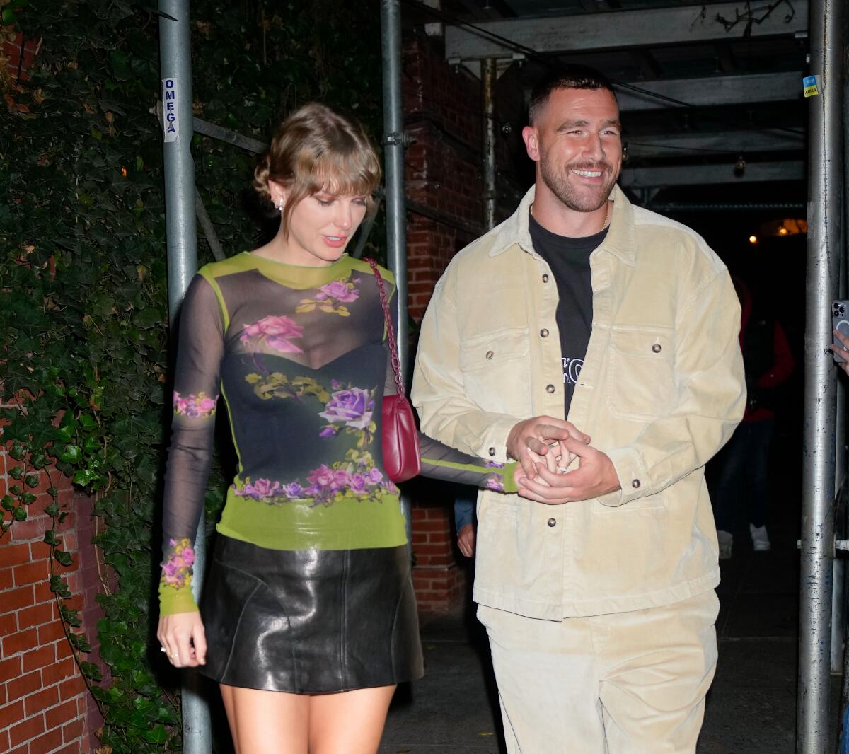 Taylor Swift in a black mini and colorful long-sleeve shirt holds hands with Travis Kelce in tan pants and a tan jacket