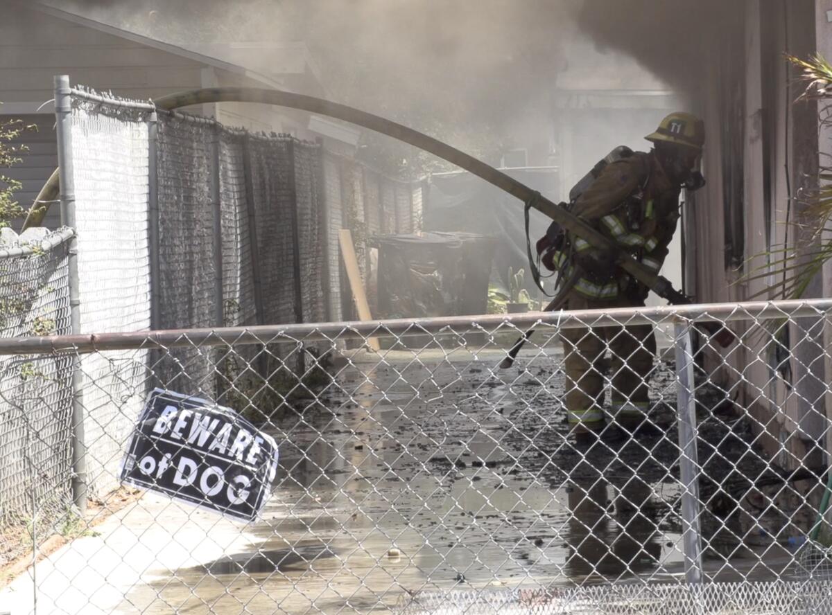 A firefighter holds a hose inside a smoky, burned-out structure behind chain-link fencing.