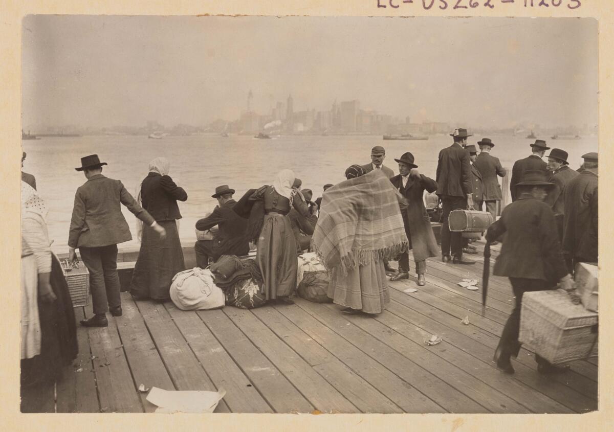 A black-and-white photo of immigrants on a dock facing the New York City skyline
