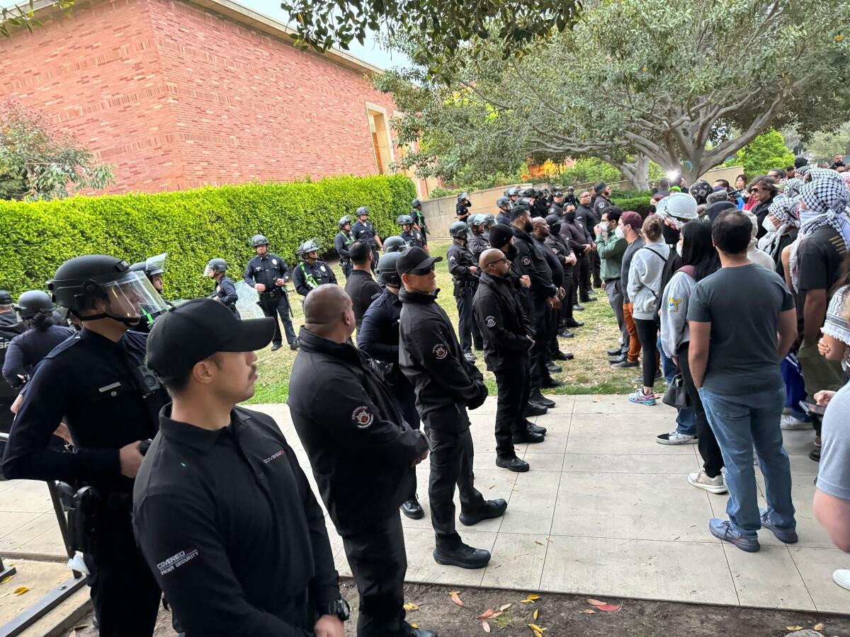 A line of men in dark uniforms, some in riot gear, face lines of protesters in a grassy area of campus.