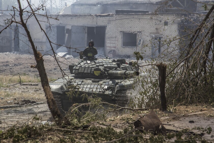 A solder fires from atop a tank in front of a destroyed building