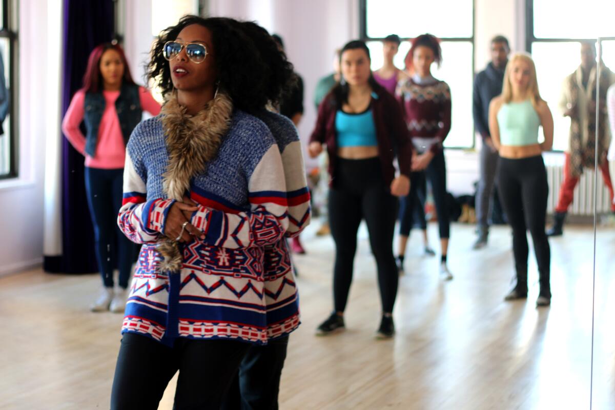 A woman stands before dancers in a rehearsal hall.