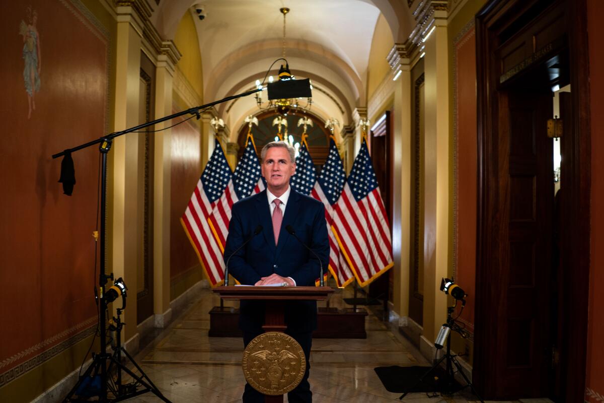 A man stands in an ornate hallway at a lectern with a bank of American flags behind him