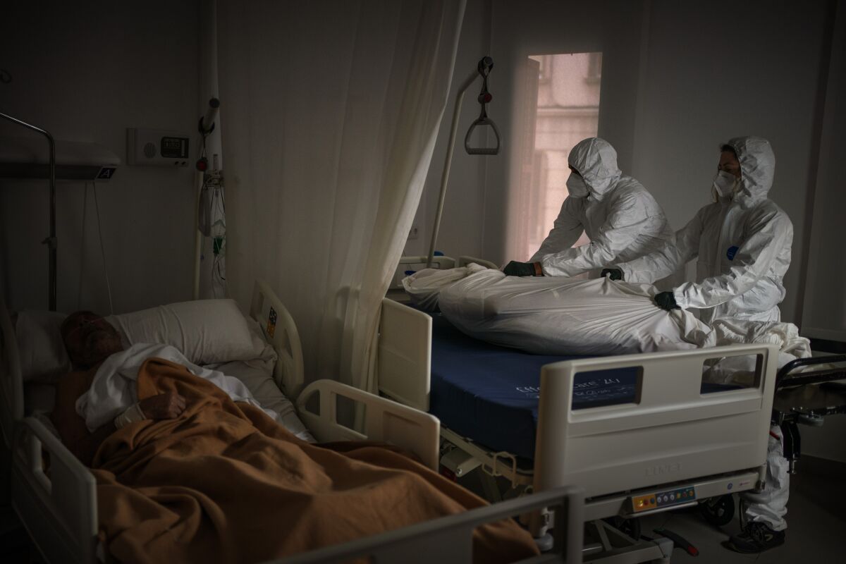 Wearing protective suits, funeral home workers remove the body of an elderly person who died of COVID-19 at a nursing home while another resident sleeps in his bed in Barcelona, Spain, Thursday, Nov. 5, 2020. In 2020, Spaniards have normalized things unimaginable only 12 months before. But 2020 will also go down as the year in which an unknown virus shook the foundations of the social contract and threw into question a system that failed to prevent so many deaths. (AP Photo/Emilio Morenatti)