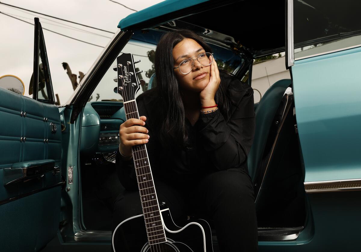 A musician sits in a car with the door open, holding a guitar.