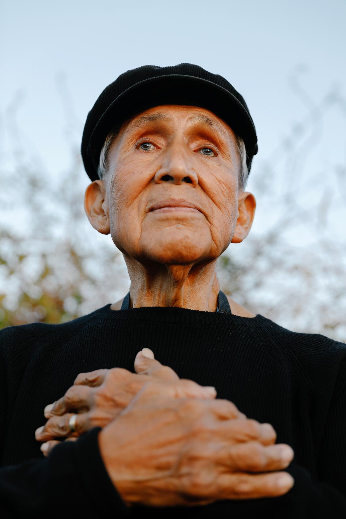 A close-up of a man dressed in black, wearing a cap and folding his hands across his chest