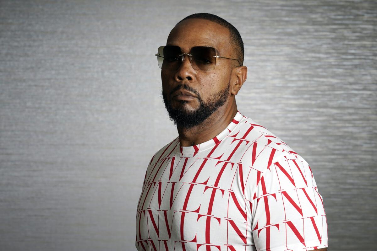 Timbaland poses for a portrait in front of a gray background clad in sunglasses and a white shirt with red letters
