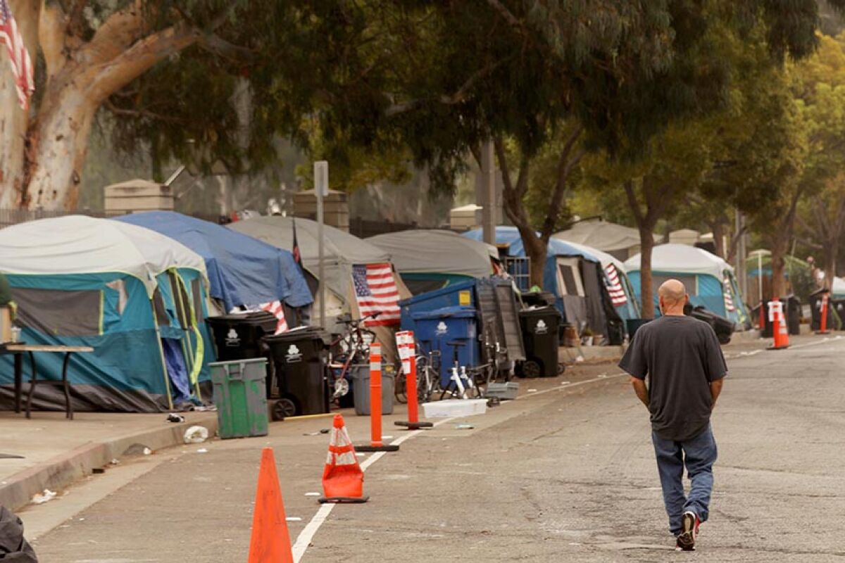 A homeless veteran walks along a street with tents on the sidewalk.