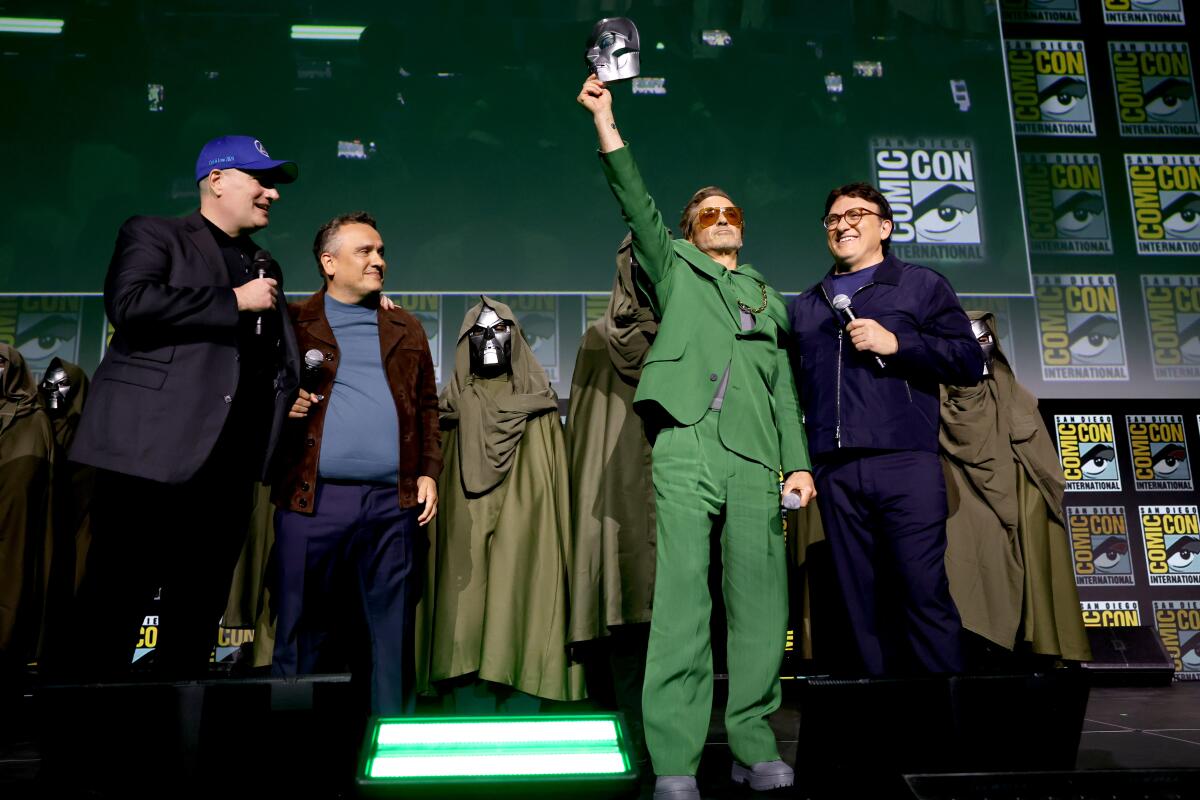 Three men looking at a man in a green suit holding up a mask on a stage surrounded by cloaked figures
