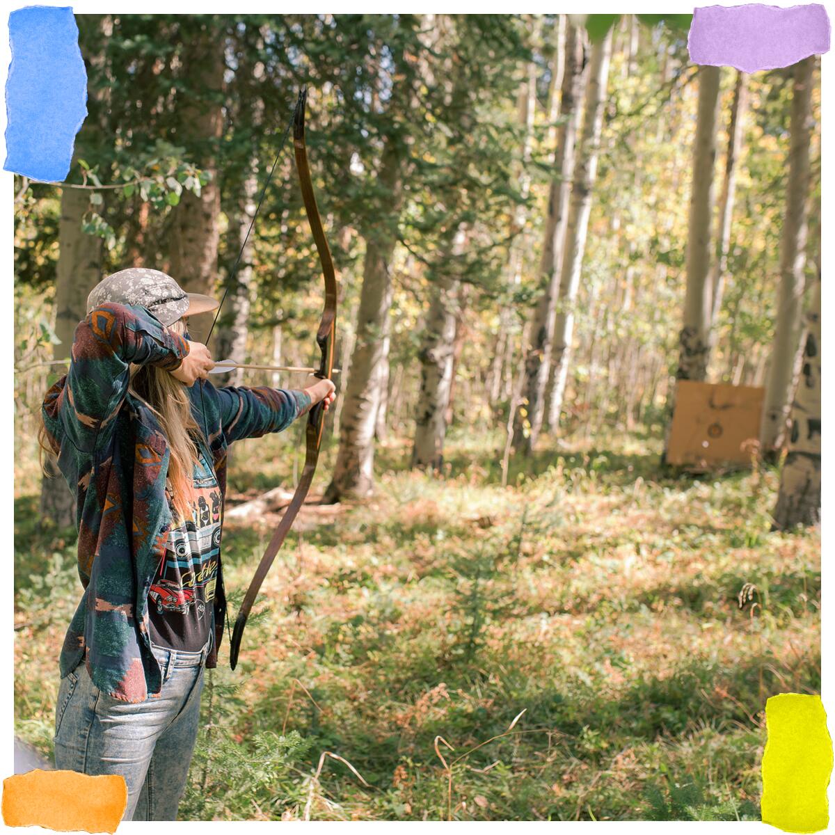 A person with long hair aims a wooden bow at a target in the woods.