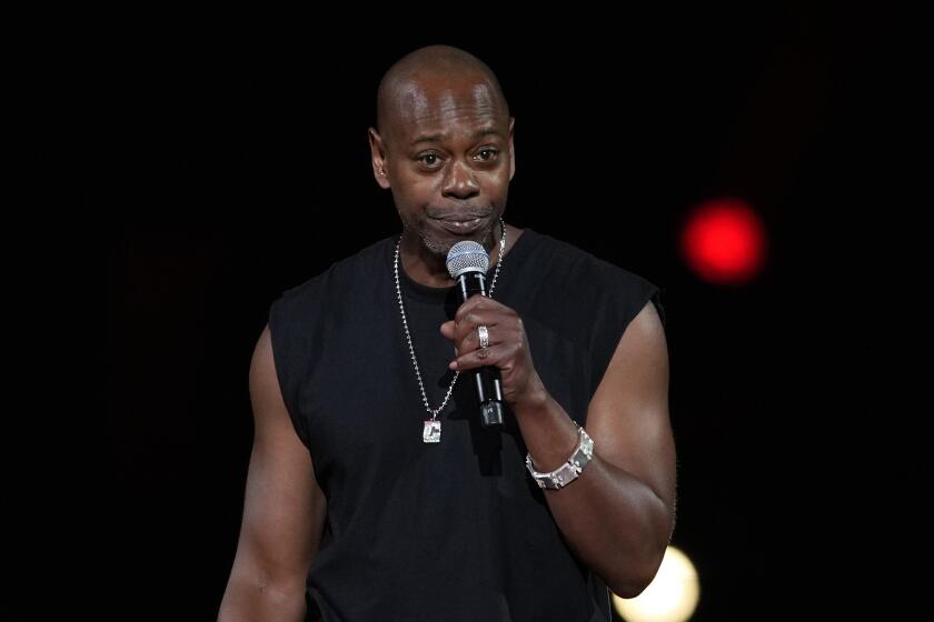 Dave Chappelle talks into microphone in a black sleeveless shirt while performing on stage