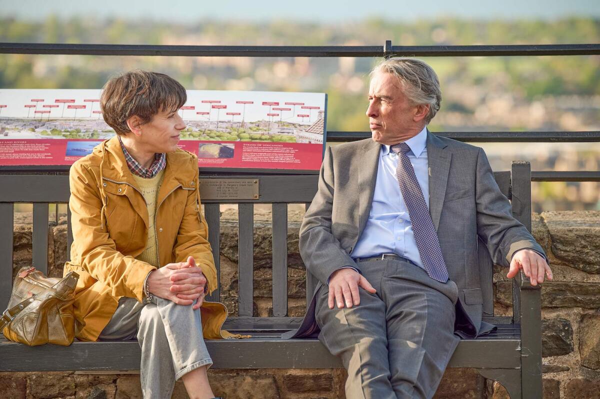 Sally Hawkins and Steve Coogan sit on a wooden bench looking at each other.