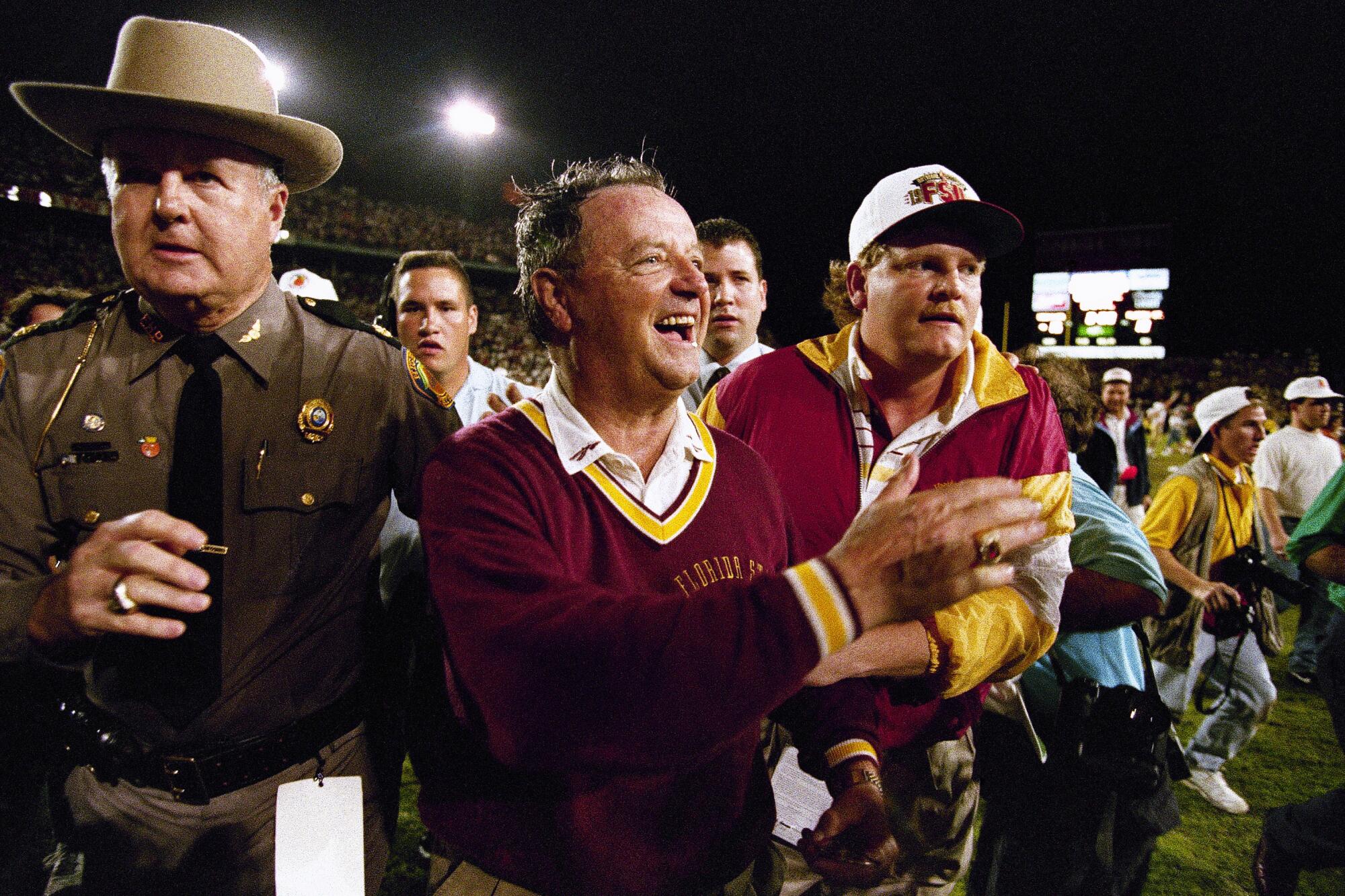 Bobby Bowden, smiling, stands with a crowd of other people on the football field.