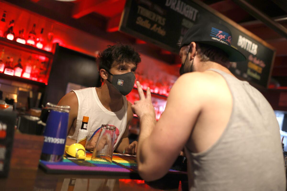 A bartender wearing a mask stands behind bar taking an order from a customer seated at the bar