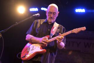 Dave Mason in a black button-down shirt playing a guitar on a stage 