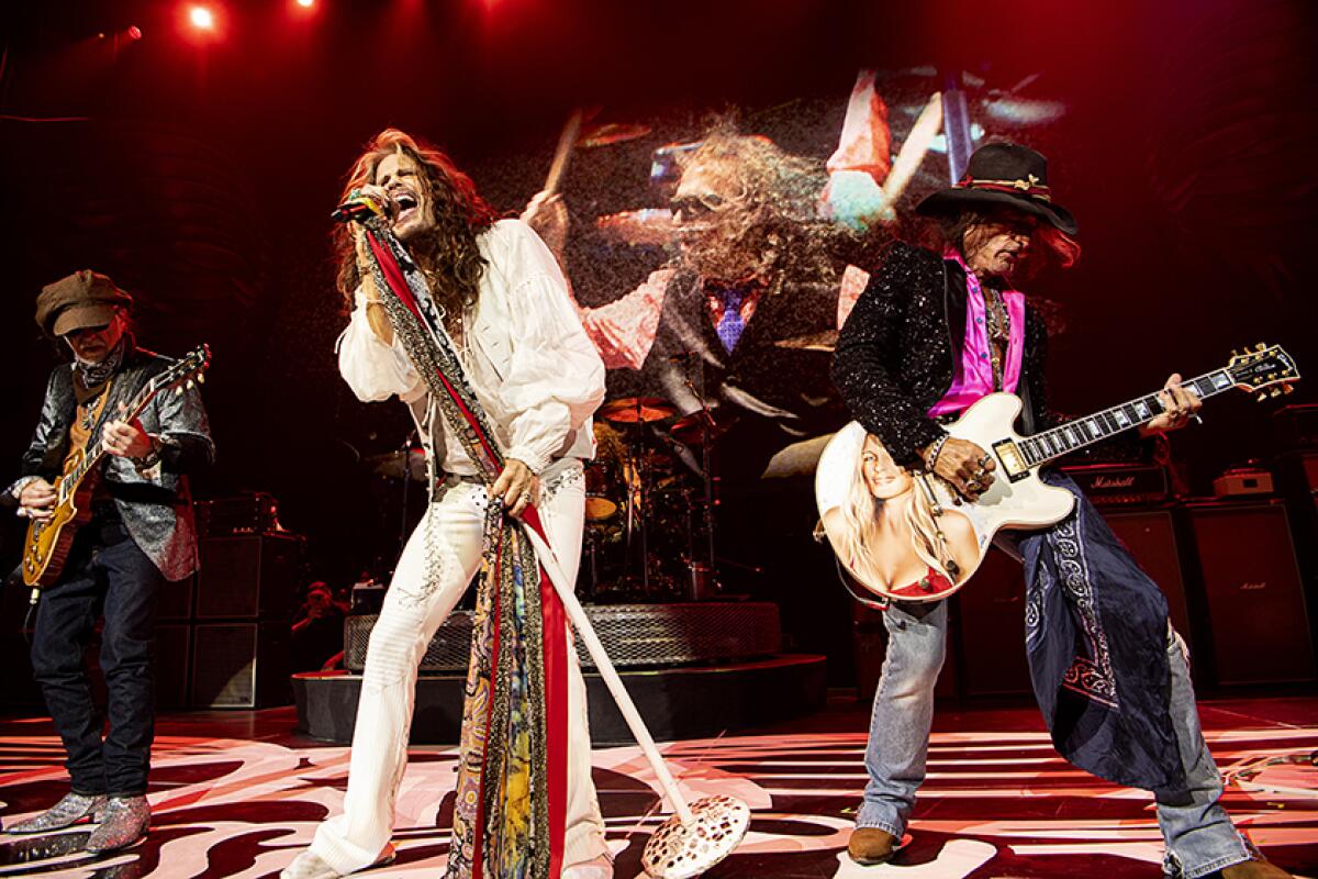 Steven Tyler wearing white sings into a microphone while members of his rock band play guitars and drums around him.