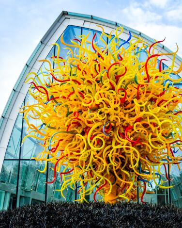 A large spiraling yellow and red glass sculpture outside an arched glasshouse wall.