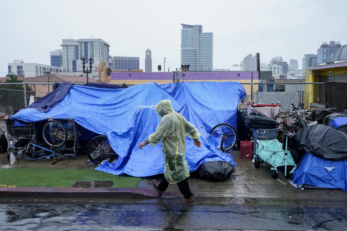 A person in a rain poncho walks in front of a street encampment.