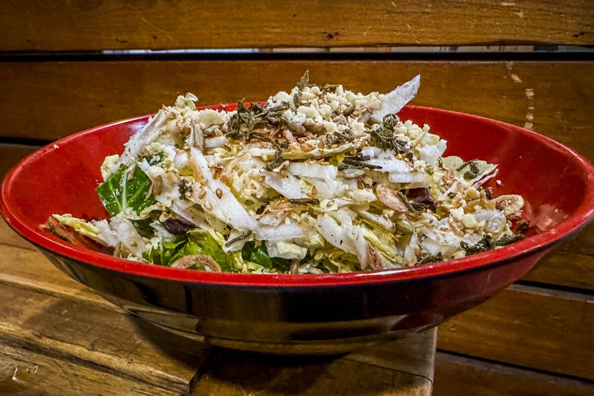 A salad of jasmine tea leaves, Napa cabbage, mixed seeds, peanuts and fried shallots in a red bowl.