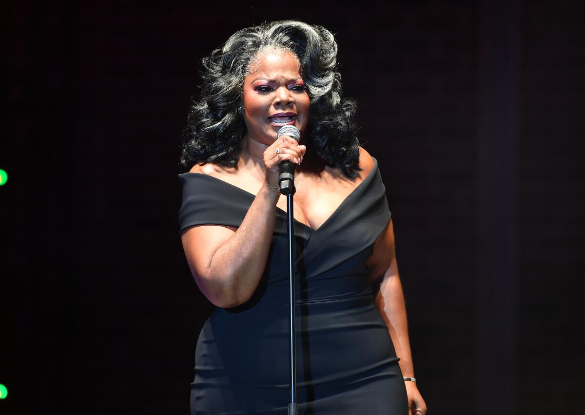 Mo'Nique holds a microphone while performing comedy onstage in a black, off-the-shoulder dress