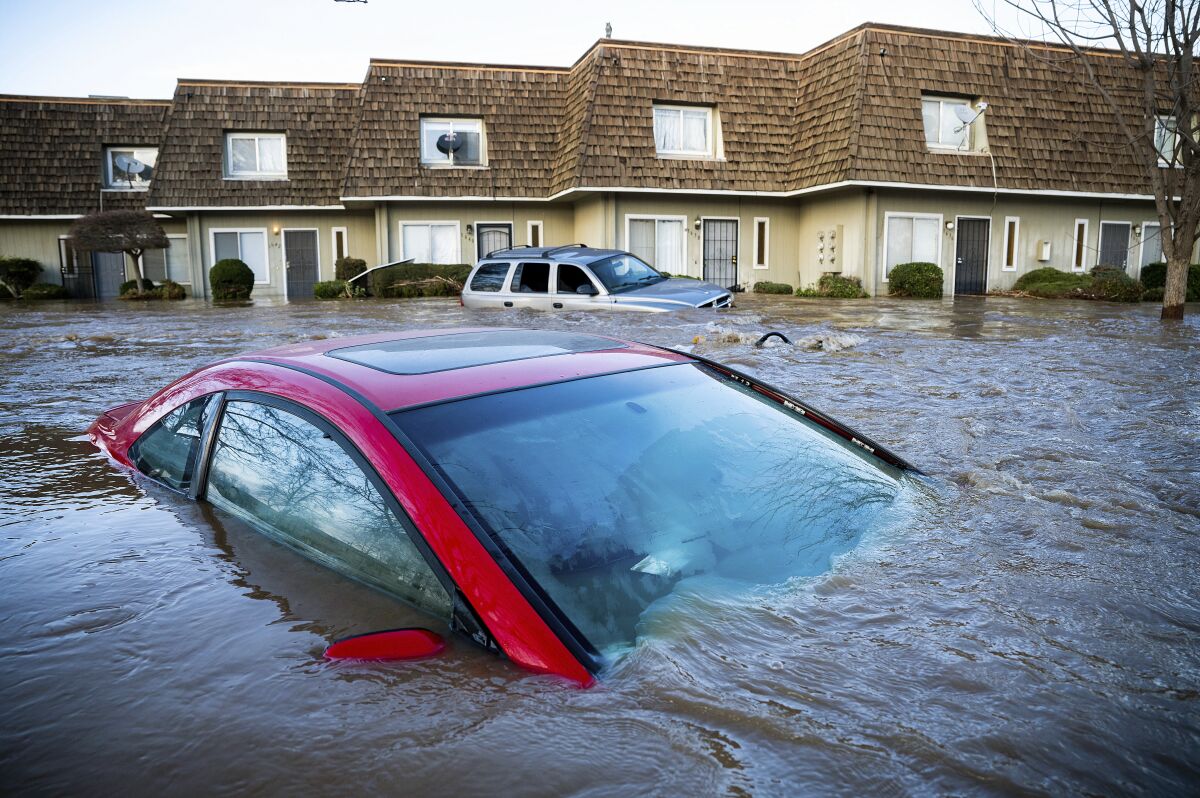 A car is submerged in floodwater up to its windows. Another flooded car and a row of townhouses are in the background. 