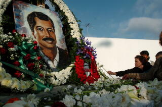 People place single blooms before a portrait of a smiling man with dark hair and mustache, surrounded by flowers and wreaths