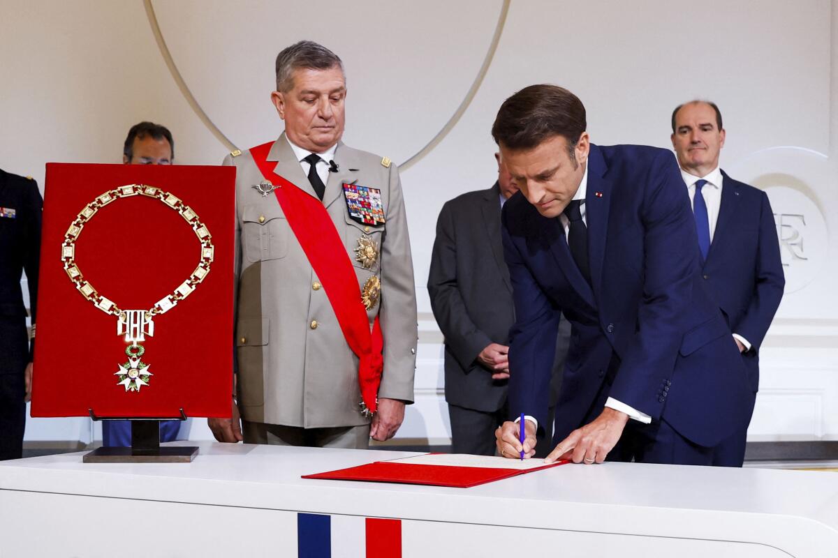 A man in military uniform stands next to a man in a blue suit who is signing a document.