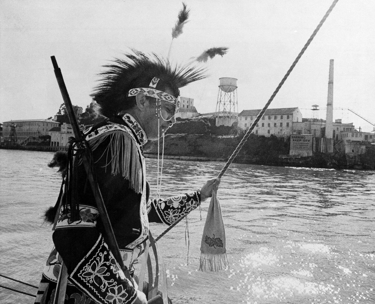 Black-and-white photo of a man in traditional Native American dress with Alcatraz in the background